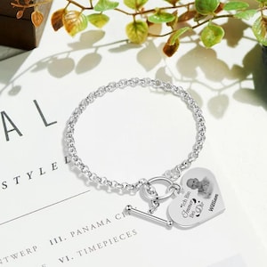 Op de afbeelding: Een zilveren armband met een hartvormige bedel. De bedel heeft een foto en de Duitse tekst "Ich Bin Immer Bei Dir" en de naam "William". De armband ligt op een wit oppervlak met groene bladeren.