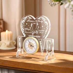 Könnte beinhalten: Eine transparente Acryluhr mit herzförmigem Aufsatz, mit den Worten "Forever & Always" und "Always Forever in my Heart". Das Zifferblatt ist goldfarben mit römischen Ziffern. Das Wort "LOVE" ist darunter eingraviert.