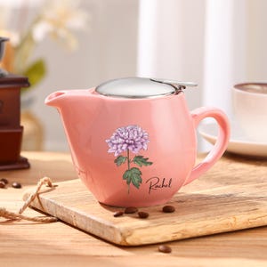 Puede incluir: Tetera de cerámica rosa con tapa y asa de acero inoxidable. La tetera presenta un diseño de flor morada y el nombre "Rachel" en escritura cursiva. La tetera está sobre una tabla de cortar de madera.