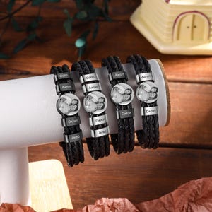 Könnte beinhalten: Vier schwarze geflochtene Lederarmbänder mit personalisierten Fotoanhängern und gravierten Namensschildern. Die Armbänder werden auf einem weißen Ständer präsentiert, der das individuelle Design zeigt. Jedes Armband hat einen anderen Namen eingraviert.