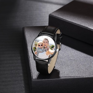Puede incluir: Reloj de pulsera negro con correa de cuero. La esfera del reloj muestra una fotograf&iacute;a en color de una mujer y un ni&ntilde;o. El reloj tiene un bisel negro y marcadores de hora plateados. El reloj est&aacute; sobre una caja negra.