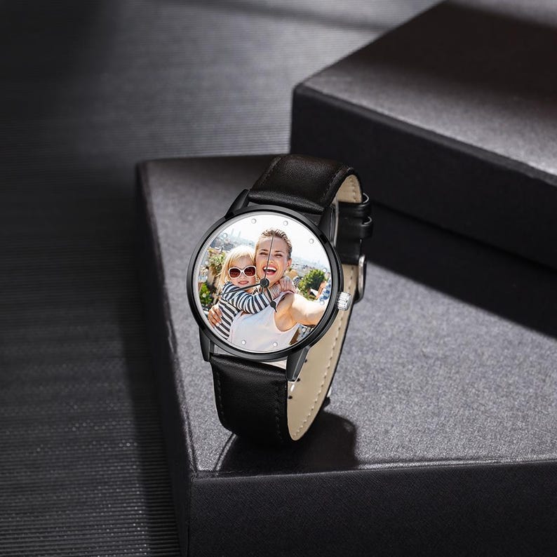 Puede incluir: Reloj de pulsera negro con correa de cuero. La esfera del reloj muestra una fotograf&iacute;a en color de una mujer y un ni&ntilde;o. El reloj tiene un bisel negro y marcadores de hora plateados. El reloj est&aacute; sobre una caja negra.