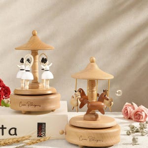 Puede incluir: Dos cajas de música de carrusel de madera. Una presenta figuras de bailarinas, la otra figuras de caballos. Ambas son de color marrón claro con un techo abovedado y una base de madera. El nombre "Cesar Rodriguez" está grabado en la base de cada caja de música.
