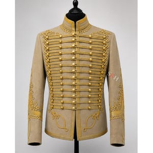 May include: A beige jacket featuring a high collar and gold-toned corded embroidery across the front. The garment includes multiple horizontal rows of gold buttons and decorative gold braiding on the sleeves and chest, with the letters SKH visible.