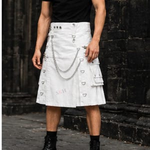 May include: A white utility kilt featuring multiple cargo pockets, silver-toned metal hardware, and a double-chain attachment draped across the front. The garment includes a high waistband with button closures and the letters SKH printed in red.