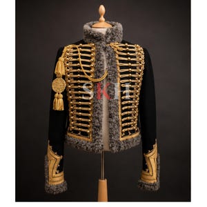 May include: Black military-style jacket with gold embellishments, including buttons, trim, and tassels. The jacket features a high collar with gray faux fur trim and gold embroidery on the sleeves. The jacket is displayed on a mannequin.