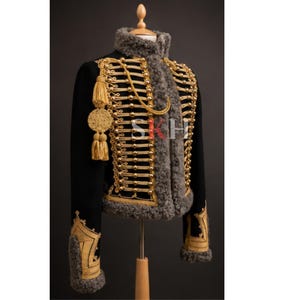 May include: A black military-style jacket featuring gold braided cord detailing, metallic buttons, and grey faux fur trim along the collar, front opening, and cuffs. The jacket includes gold tassels and the letters SKH printed on the front.