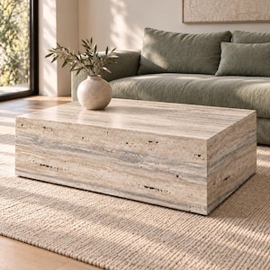 May include: A rectangular coffee table crafted from beige travertine stone with a porous, natural texture. The table features a low-profile, block-like design with visible horizontal veining and a smooth top surface holding a round vase.