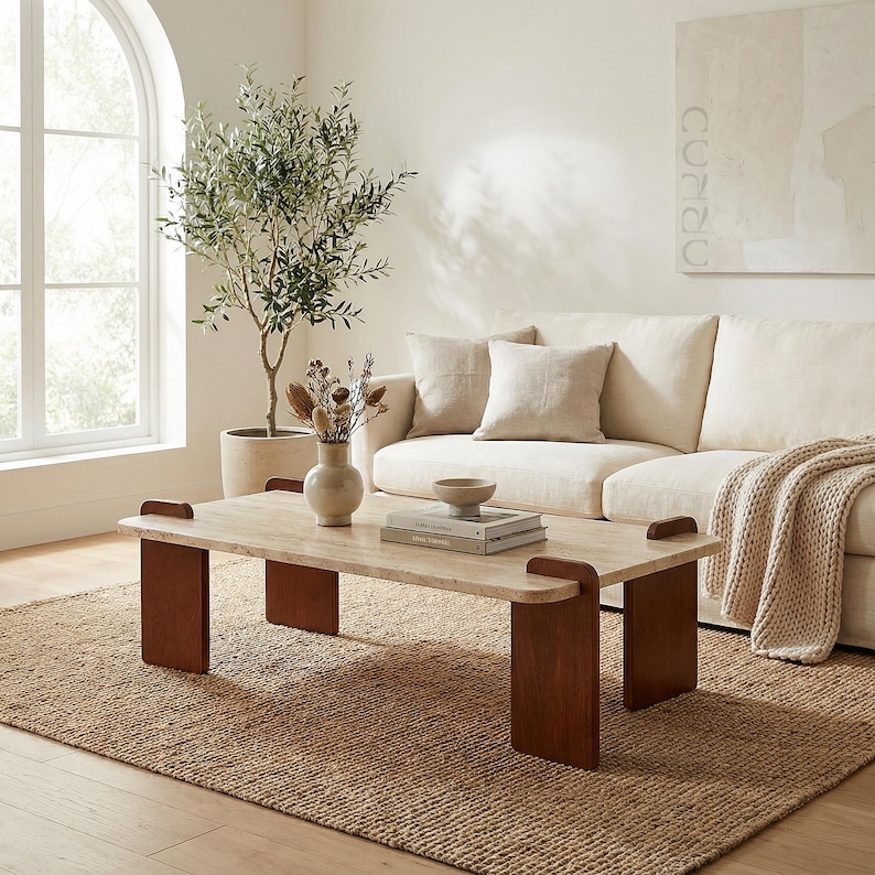 May include: A rectangular coffee table featuring a light beige stone top with rounded corners and four dark brown wooden slab legs. The table is positioned on a textured jute rug, holding a small ceramic vase with dried botanicals and books.