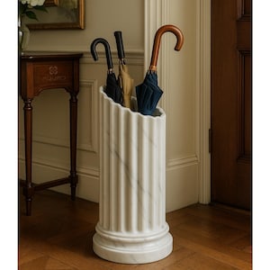 May include: A white marble umbrella stand with a fluted, column-like design. The cylindrical stand features vertical grooves and a circular base, holding three folded umbrellas with black, tan, and navy blue fabric and curved handles.