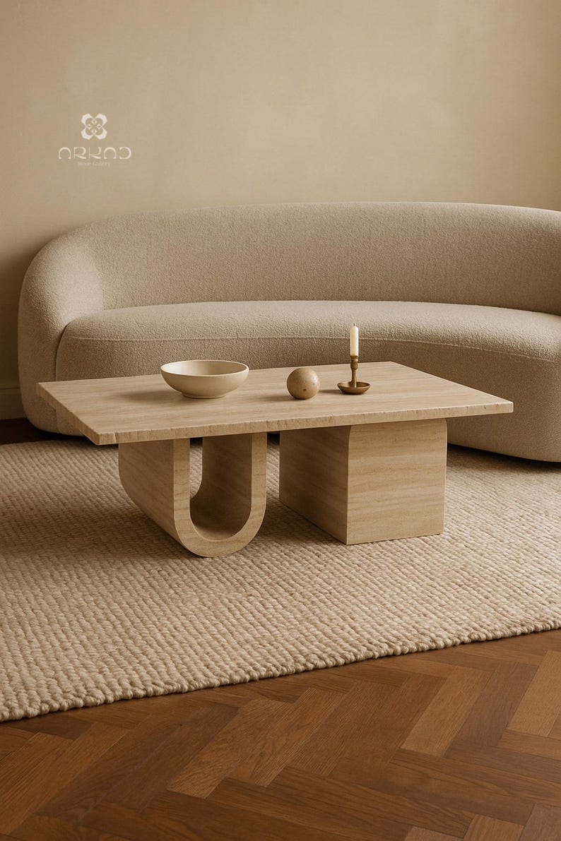 May include: A light wood rectangular coffee table with a distinctive U-shaped leg design. A small bowl, sphere, and candle holder with a lit candle are on the table. The table is in front of a beige sofa and sits on a woven rug.