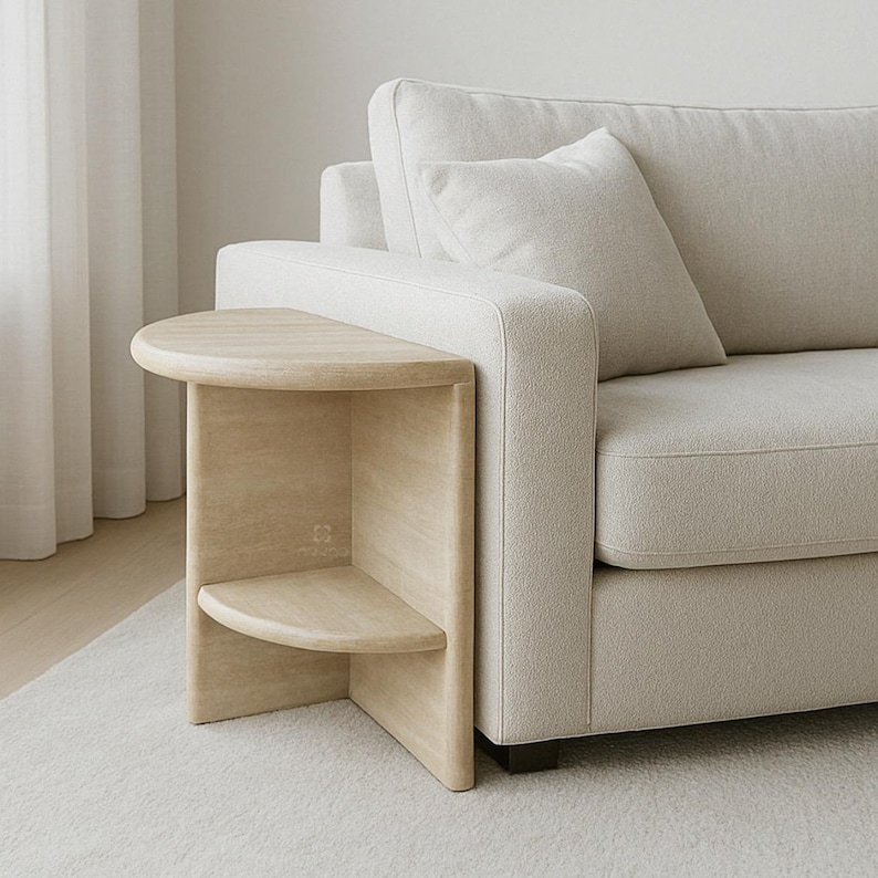 May include: A light-toned wooden side table with a semi-circular top and a matching lower shelf, positioned directly against the side of a cream-colored upholstered sofa. The table features a curved vertical support structure.