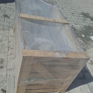 May include: A large, rectangular wooden crate wrapped in clear plastic. The crate is made from light-coloured wood and appears to be new. The plastic wrap is tightly secured around the crate, protecting its contents. The crate is on a grey, textured surface.
