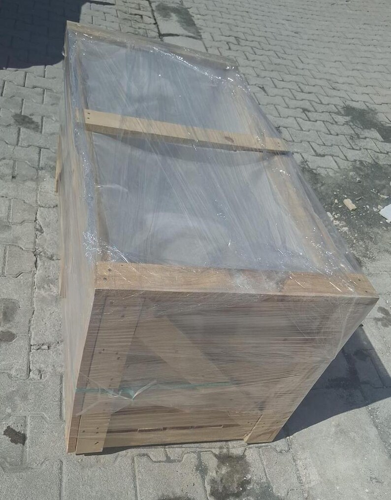 May include: A large, rectangular wooden crate wrapped in clear plastic. The crate is made from light-coloured wood and appears to be new. The plastic wrap is tightly secured around the crate, protecting its contents. The crate is on a grey, textured surface.