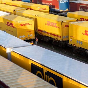 May include: An elevated shot of a train yard with numerous yellow DHL shipping containers. The containers, branded with the DHL logo, are loaded onto train carriages. Other carriages are visible, some with red and blue containers in the background.