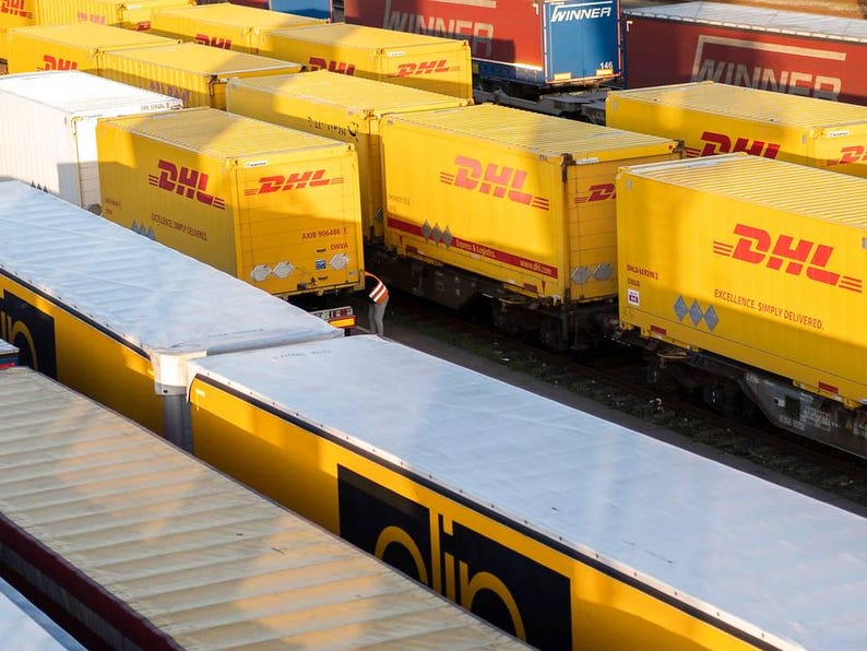 May include: An elevated shot of a train yard with numerous yellow DHL shipping containers. The containers, branded with the DHL logo, are loaded onto train carriages. Other carriages are visible, some with red and blue containers in the background.