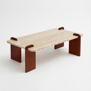 May include: A rectangular coffee table featuring a beige, porous travertine stone top with rounded corners. The tabletop is supported by four dark brown wooden legs that slot into the edges of the stone surface, creating a visible joint.