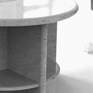 May include: A semi-circular side table crafted from light-colored stone with a porous, speckled texture. The table features a smooth, rounded top surface supported by a central vertical pillar and a lower shelf, showcasing a natural finish.