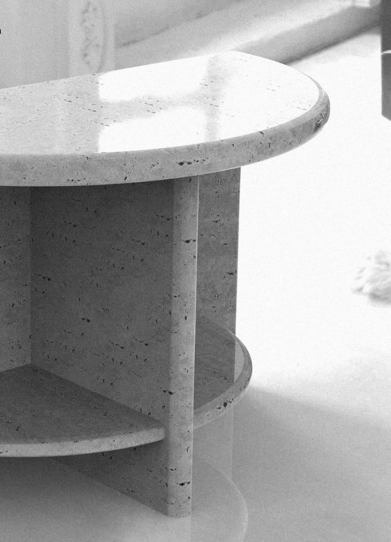 May include: A semi-circular side table crafted from light-colored stone with a porous, speckled texture. The table features a smooth, rounded top surface supported by a central vertical pillar and a lower shelf, showcasing a natural finish.