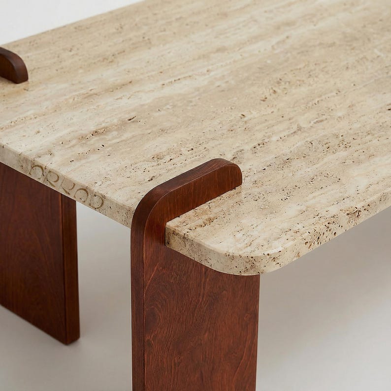 May include: A rectangular table featuring a thick, porous beige travertine stone top with rounded corners. The tabletop is supported by two dark brown wooden legs that curve over the edges of the stone, securing it in place.