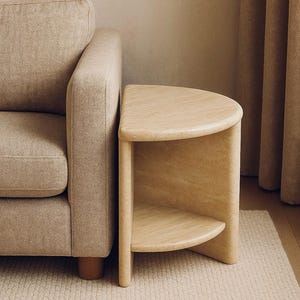 May include: A beige stone side table with a semi-circular top and a matching lower shelf. The table features the text Halo side table, Arkad stone gallery, 2025 - COLLECTION. It is positioned next to a beige upholstered sofa.