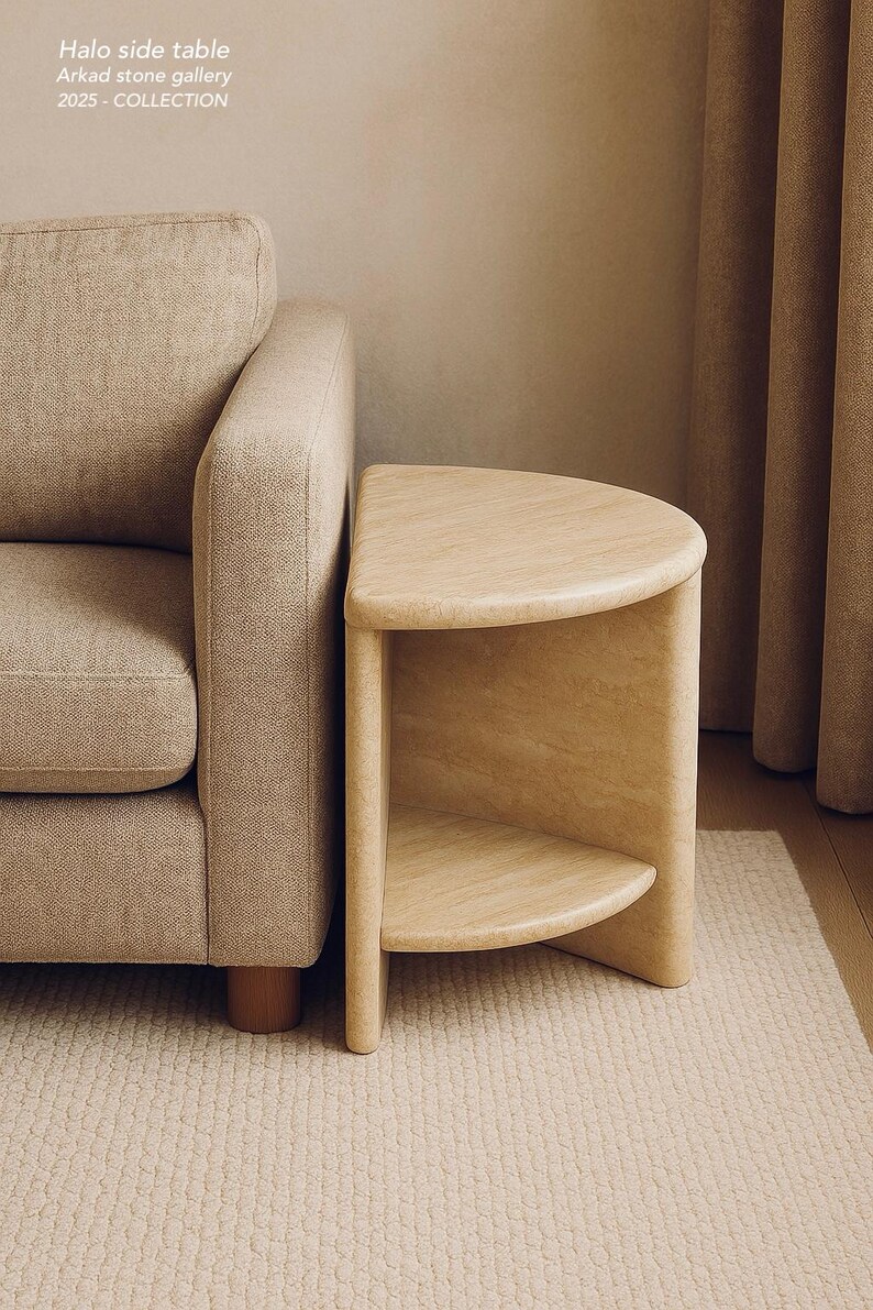 May include: A beige stone side table with a semi-circular top and a matching lower shelf. The table features the text Halo side table, Arkad stone gallery, 2025 - COLLECTION. It is positioned next to a beige upholstered sofa.