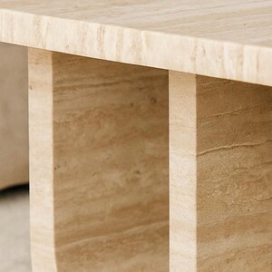 May include: Close-up of a light beige stone table. The table has a rectangular top and two U-shaped legs. The stone has a natural, textured appearance with variations in colour.