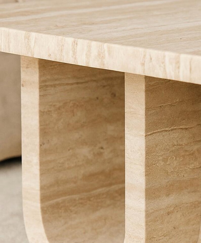 May include: Close-up of a light beige stone table. The table has a rectangular top and two U-shaped legs. The stone has a natural, textured appearance with variations in colour.