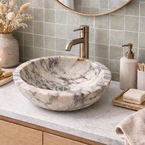 May include: A marble washbasin with a grey and beige pattern sits on a white countertop. A brushed nickel tap is mounted above the basin. A soap dispenser, a tray with folded towels, and a vase with dried flowers are also visible.