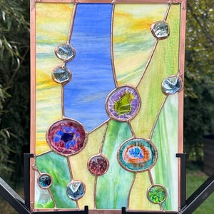 May include: A rectangular stained glass panel framed in copper, featuring a vertical composition of yellow, blue, and green glass segments accented by various small, circular, and multi-colored glass cabochons soldered onto the surface.