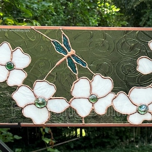 May include: A stained glass window panel featuring white dogwood blossoms with green centers and a blue dragonfly in the center.