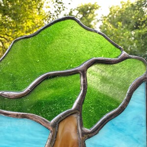 May include: A stained glass panel featuring a tree design with vibrant green glass for the canopy, a brown glass trunk, and light blue glass for the sky. The pieces are joined by dark metal solder lines, creating a clear outline of the tree shape.