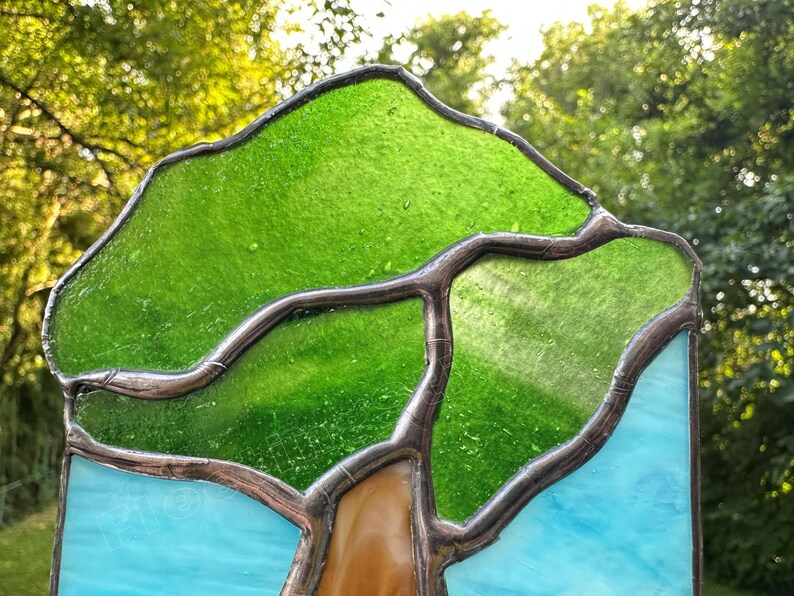 May include: A stained glass panel featuring a tree design with vibrant green glass for the canopy, a brown glass trunk, and light blue glass for the sky. The pieces are joined by dark metal solder lines, creating a clear outline of the tree shape.