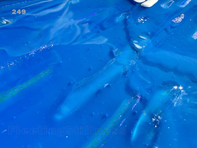 May include: A vibrant blue liquid surface with a glossy, reflective texture. The number 249 is printed in white in the upper left corner, and the text FleetingStillness is printed in white along the bottom edge of the frame.