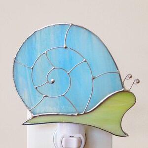 May include: A blue and green stained glass nightlight shaped like a snail. The snail has a light blue shell with a white swirl pattern and a green body.