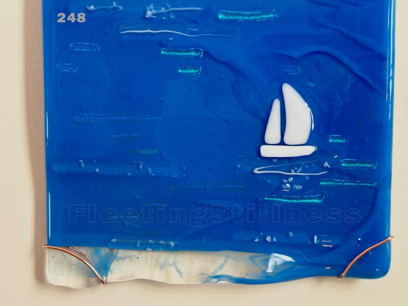 May include: A rectangular fused glass art piece featuring a deep blue background with a white sailboat silhouette. The text 248 appears in the top left corner, and the text FleetingStillness is centered near the bottom. Copper wire accents the base.