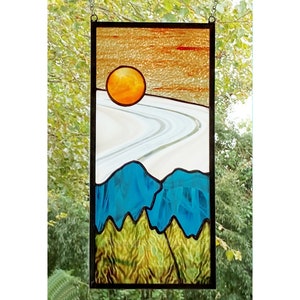 May include: A stained glass window depicting a stylized landscape with a setting sun, a winding road, and blue mountains. The sun is a bright orange, the road is white with a gray outline, and the mountains are a deep blue with green trees at the base.