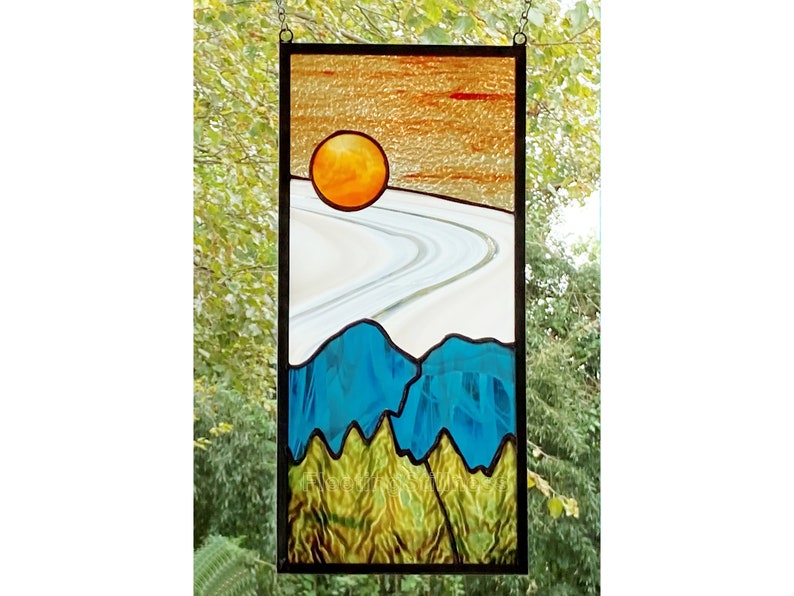 May include: A stained glass window depicting a stylized landscape with a setting sun, a winding road, and blue mountains. The sun is a bright orange, the road is white with a gray outline, and the mountains are a deep blue with green trees at the base.