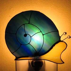 May include: A stained glass nightlight shaped like a snail with a blue and green shell and a yellow body. The nightlight is plugged into an electrical outlet.