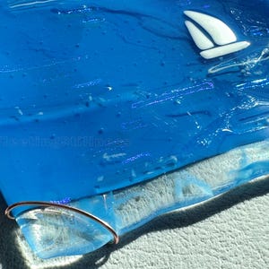 May include: A square blue fused glass plate featuring a white sailboat graphic in the upper right corner. The number 248 is printed in the top left, and the text FleetingStillness is visible. A thin copper wire loop is attached to the bottom edge.