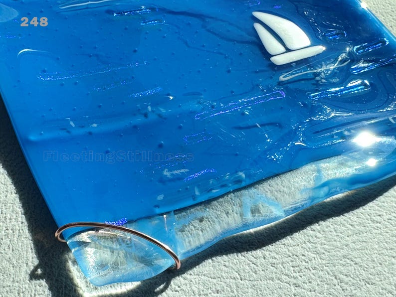 May include: A square blue fused glass plate featuring a white sailboat graphic in the upper right corner. The number 248 is printed in the top left, and the text FleetingStillness is visible. A thin copper wire loop is attached to the bottom edge.