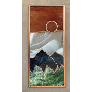 May include: A stained glass window depicting a sunset over mountains. The sky is a gradient of orange, white, and blue. The mountains are green and black. The sun is a circle of orange glass.
