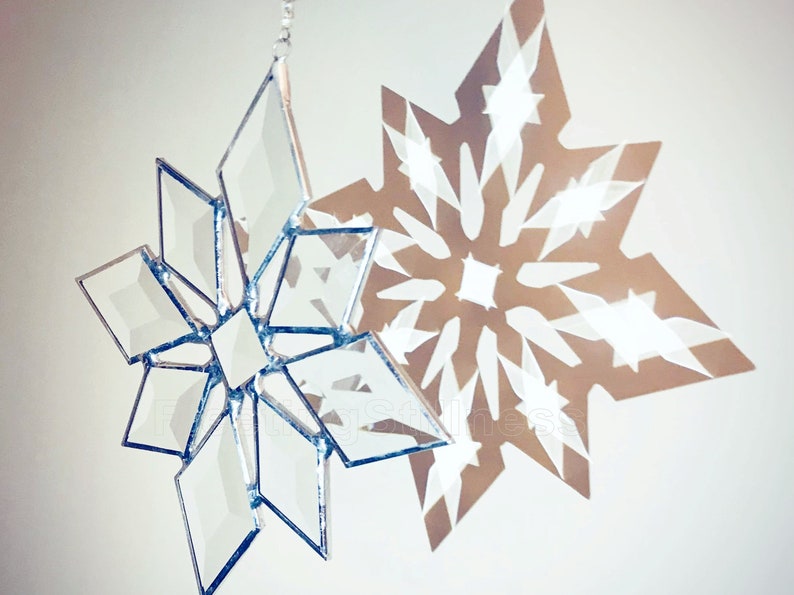 May include: A silver glass snowflake ornament hangs from a string, casting a shadow of a white snowflake on a white wall.