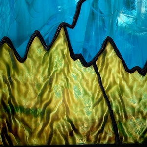 May include: A stained glass window panel depicting a mountain range with a blue sky above. The mountains are outlined in black and have a textured, green surface.