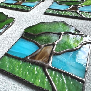 May include: A set of square stained glass panels featuring a tree design with a brown trunk, green foliage, and a bright blue sky background. Each panel is constructed with dark metal solder lines separating the individual glass pieces.