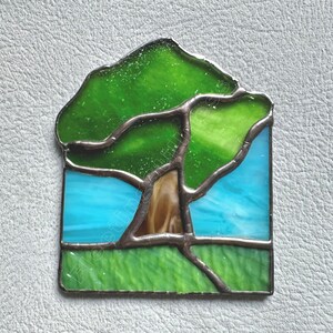 May include: A stained glass suncatcher featuring a tree with a brown trunk and green canopy set against a bright blue sky and a green grassy foreground. The individual glass pieces are joined by dark metal solder lines in a landscape design.