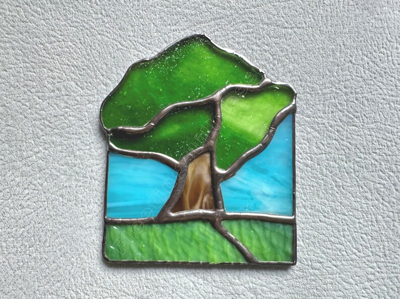May include: A stained glass suncatcher featuring a tree with a brown trunk and green canopy set against a bright blue sky and a green grassy foreground. The individual glass pieces are joined by dark metal solder lines in a landscape design.