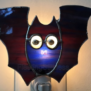 May include: A stained glass bat night light with a blue and red body and large white eyes. The night light is plugged into an outlet.