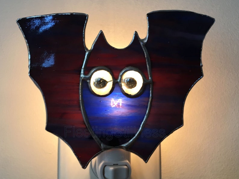 May include: A stained glass bat night light with a blue and red body and large white eyes. The night light is plugged into an outlet.