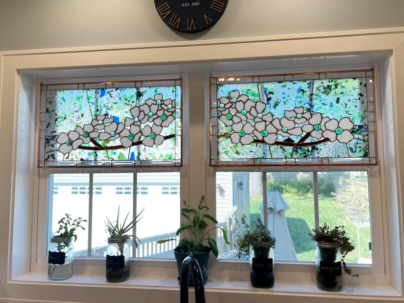 May include: Two rectangular stained glass window panels featuring white dogwood flowers with green glass centers on brown branches, set against a background of clear, blue, and green textured glass pieces held together by copper foil solder lines.